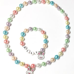 May include: A pastel colored beaded necklace and bracelet set with bunny charms. The necklace has a charm with a bunny face and the bracelet has a charm with the name "ALLI" on it.
