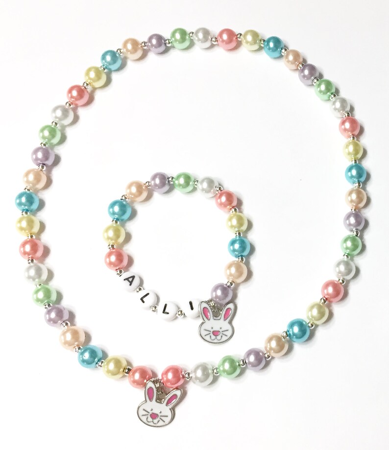 May include: A pastel colored beaded necklace and bracelet set with bunny charms. The necklace has a charm with a bunny face and the bracelet has a charm with the name "ALLI" on it.