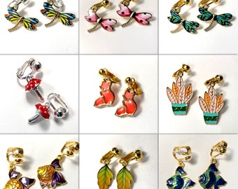 May include: Assortment of colorful clip-on earrings featuring various designs, including dragonflies, mushrooms, foxes, plants, and fish. The earrings are made of metal and have a gold finish.