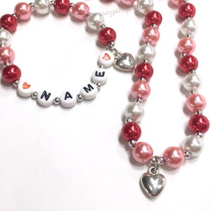 May include: A red, white and pink beaded necklace and bracelet set with silver heart charms. The bracelet has white beads with the word "NAME" spelled out in black letters.