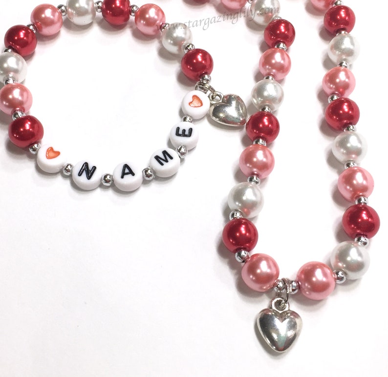 May include: A red, white and pink beaded necklace and bracelet set with silver heart charms. The bracelet has white beads with the word "NAME" spelled out in black letters.
