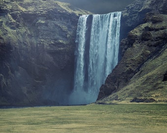 Op de afbeelding: Een grote waterval stort naar beneden van een rotsachtige klif, met een groen grasveld op de voorgrond. De lucht is bewolkt met een vleugje zonlicht.