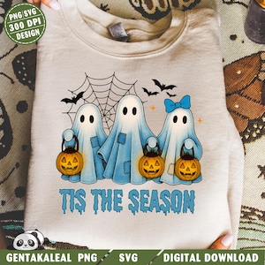 Op de afbeelding: Een lichtbeige sweatshirt met een Halloween-ontwerp. Het ontwerp bevat drie cartoongeesten met pompoenlantaarns, een spinnenweb, vleermuizen en de tekst "TIS THE SEASON" in blauw. De geesten hebben blauwe pleisters en één heeft een blauwe strik.