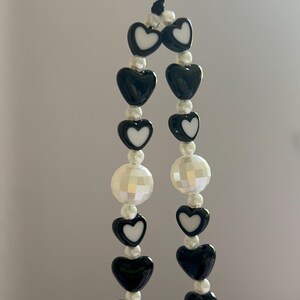 May include: Black and white beaded phone charm. The charm features alternating black heart-shaped beads with white centers, and faceted white beads. The charm is strung on a black cord.