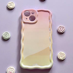 May include: A pastel-colored phone case with a wavy edge, featuring a gradient of pink, peach, and yellow. Scattered around the case are heart-shaped candies with various phrases like "Be Kind" and "Good Vibes".
