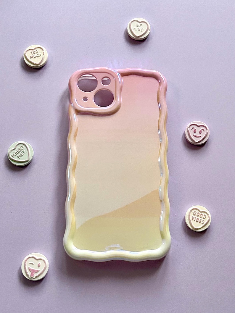 May include: A pastel-colored phone case with a wavy edge, featuring a gradient of pink, peach, and yellow. Scattered around the case are heart-shaped candies with various phrases like "Be Kind" and "Good Vibes".