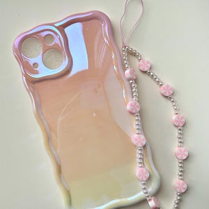 May include: Iridescent pink phone case with a wavy edge, next to a beaded phone strap. The strap features alternating pink and white candy-shaped beads and silver-colored beads. The phone case has cutouts for the camera and other phone features.