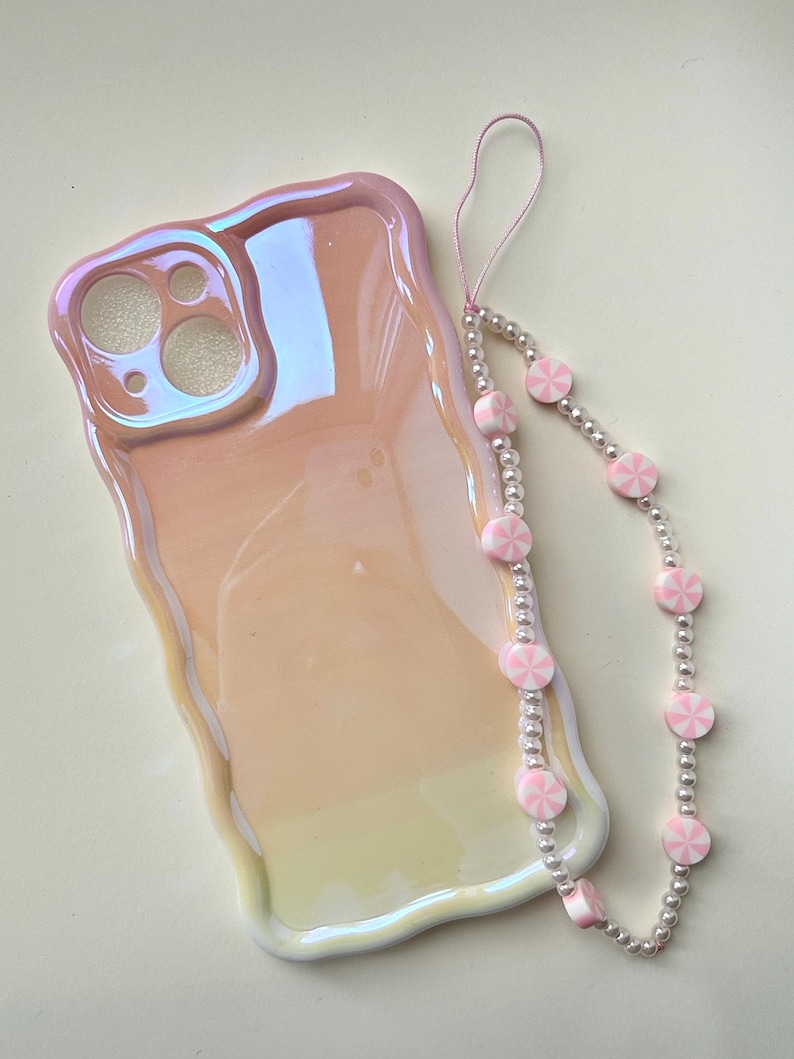 May include: Iridescent pink phone case with a wavy edge, next to a beaded phone strap. The strap features alternating pink and white candy-shaped beads and silver-colored beads. The phone case has cutouts for the camera and other phone features.