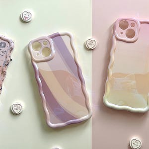 May include: Three pastel-colored phone cases with wavy edges are displayed on a light pink and green background. One case has a celestial pattern, another has a rainbow design, and the third is a gradient of pink and yellow.