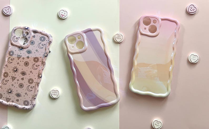 May include: Three pastel-colored phone cases with wavy edges are displayed on a light pink and green background. One case has a celestial pattern, another has a rainbow design, and the third is a gradient of pink and yellow.