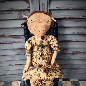 May include: A fabric doll with brown yarn hair and a hand-stitched face, wearing a cream-colored dress featuring a green and red holly leaf pattern. The doll has striped legs and is adorned with two small jingle bells on the bodice of the dress.