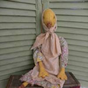 May include: A handmade yellow fabric doll with a floral print dress, a pink apron, and a pink scarf. The doll has long, thin legs and is sitting on a wooden box.