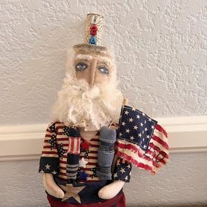 May include: A fabric doll featuring a bearded figure wearing a top hat with the letters USA, dressed in a shirt and pants patterned with American flag stars and stripes, holding a small American flag with the number 50 written on the fabric.