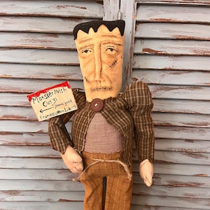 May include: A fabric doll resembling Frankenstein's monster with black hair, stitched facial details, and a brown plaid jacket over a red and white checkered shirt. It holds a sign reading Monster Mash Oct 31, graveyard, Frankenstein Lab.