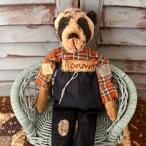 May include: A stuffed bear toy with a distressed tan and black face, wearing an orange and blue plaid shirt under dark blue denim overalls. A burlap patch on the chest features the handwritten name Roosevelt. The bear has patches on its arms and legs.