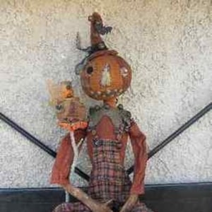 May include: A handmade rag doll pumpkin with a plaid shirt and overalls. The doll has a pumpkin head with a painted face and a black hat with a small black cat on top. The doll is holding a small pumpkin in its hand.