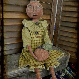 May include: A handmade rag doll dressed in a green and brown polka dot dress with a ruffled collar. The doll is holding a small orange pumpkin in its hands. The doll is sitting on a wooden bench with a rustic background.
