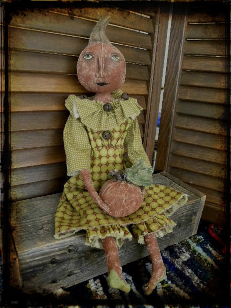 May include: A handmade rag doll dressed in a green and brown polka dot dress with a ruffled collar. The doll is holding a small orange pumpkin in its hands. The doll is sitting on a wooden bench with a rustic background.