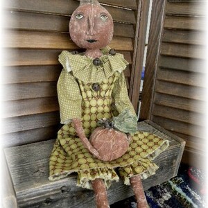 May include: A handmade rag doll dressed in a green and brown polka dot dress with a ruffled collar. The doll is holding a small brown pumpkin and is sitting on a wooden bench.