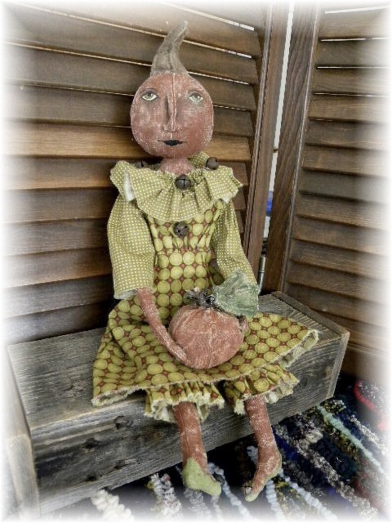 May include: A handmade rag doll dressed in a green and brown polka dot dress with a ruffled collar. The doll is holding a small brown pumpkin and is sitting on a wooden bench.