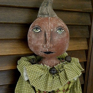 May include: A handmade fabric pumpkin doll with a green and white polka dot dress and brown bells. The doll has a painted face with brown eyes and a black mouth.