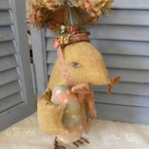 May include: A handmade fabric Easter bunny with a large, colorful hat and a pink and blue egg in its arms.