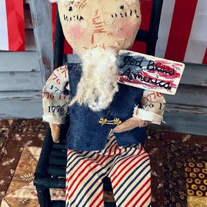 May include: A patriotic doll figure of a man in a blue vest, striped pants, and a top hat. The doll is holding a sign that reads "God Bless America." The figure is seated on a black chair, with a red and white striped background.