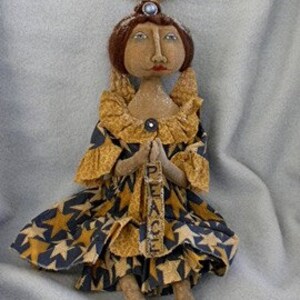May include: A handmade rag doll angel with a brown face, brown hair, and a blue dress with gold stars. The angel is holding a sign that says "Peace".