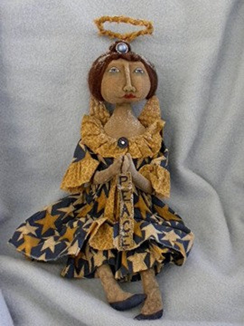 May include: A handmade rag doll angel with a brown face, brown hair, and a blue dress with gold stars. The angel is holding a sign that says "Peace".