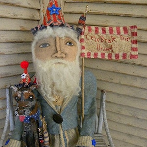 May include: A handmade rag doll dressed in blue and white striped clothing with a red, white, and blue star-shaped hat. The doll is holding a sign that says "Celebrate the Brave".