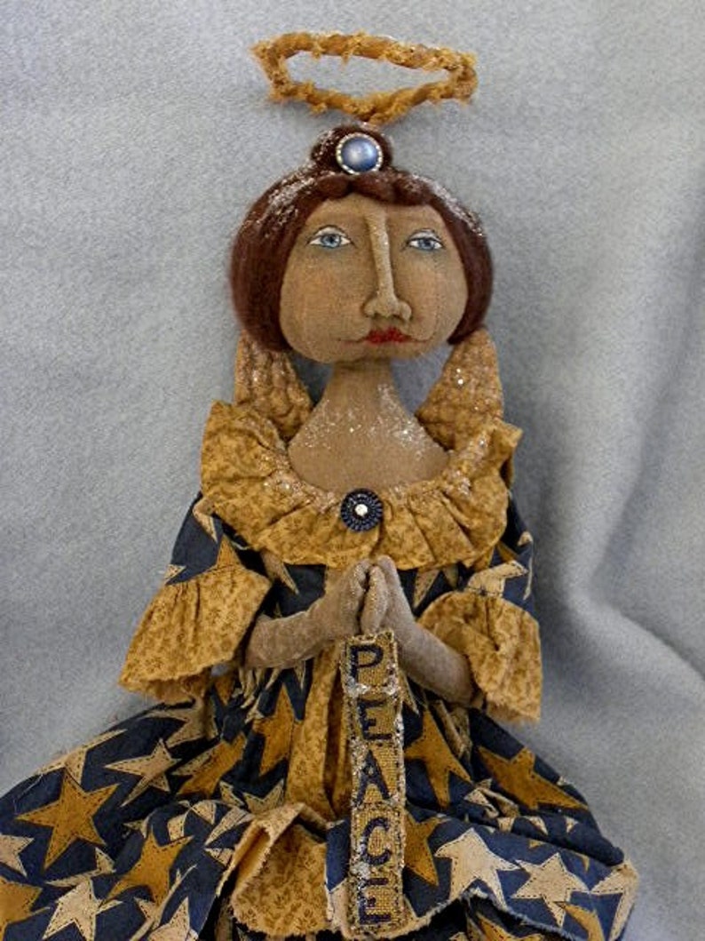 May include: A handmade rag doll angel with a halo, wearing a gold and blue dress with stars and holding a sign that says "Peace".