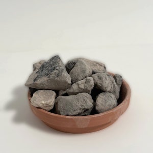 May include: A terracotta bowl filled with various gray and tan rocks. The rocks have rough textures and varying sizes. The bowl is round and has a simple, unglazed finish. The rocks appear to be natural stones.