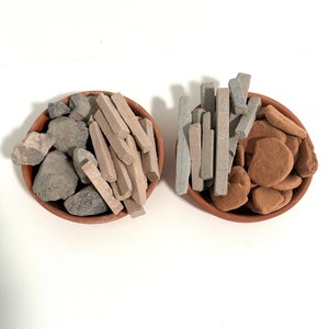 May include: Two terracotta bowls filled with natural stones. One bowl contains gray rocks and tan rectangular stones. The other bowl holds gray rectangular stones and reddish-brown, rounded stones. The bowls sit on a white surface.
