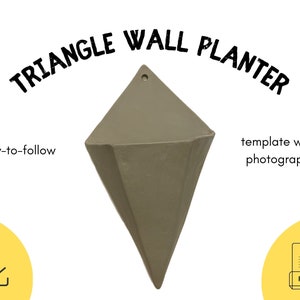 Puede incluir: Un macetero de pared triangular de color gris apagado. El macetero tiene una forma geométrica con un agujero en la parte superior para colgarlo. El texto de la imagen dice "TRIANGLE WALL PLANTER", "easy-to-follow" y "template with photographs".