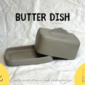 May include: A gray butter dish with a lid, sitting on a white cloth. The lid has a small handle. The words "BUTTER DISH" are printed in black above the dish. Below the dish, the text reads "with instructions and photographs."