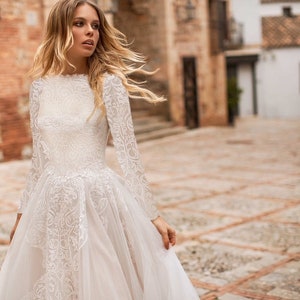 May include: A long-sleeved, floor-length white lace wedding dress with a high neck and a fitted bodice. The skirt is made of tulle and has a slight A-line shape. The dress features intricate lace detailing throughout. The logo 'Unique Modest Bridal' is visible in the bottom left corner.