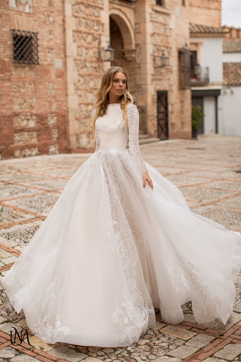 May include: A long-sleeved, ivory wedding gown with a high neckline and a full tulle skirt. The bodice features intricate lace detailing, and the skirt has delicate lace appliques along the sides. The dress is shown on a model in an outdoor setting.