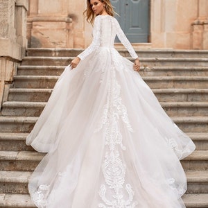 May include: A long-sleeved, floor-length wedding gown with a tulle skirt and delicate lace detailing along the back. The bodice is fitted, and the skirt flows into a train. The lace pattern is intricate and features floral motifs. The dress is ivory or off-white.  The image shows the back of the dress.