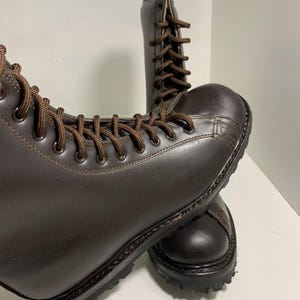May include: A pair of brown leather boots with brown laces. The boots have a rounded toe and a thick, textured sole. The boots are laced up and appear to be new.