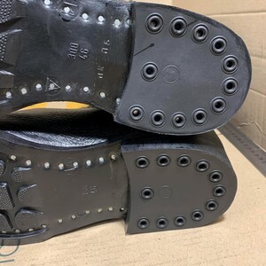 Puede incluir: Primer plano de las suelas de dos botas de cuero negro. Las suelas est&aacute;n fuertemente tachonadas con remaches met&aacute;licos y tienen un tac&oacute;n de goma grueso y duradero. Los talones tienen una serie de agujeros circulares.