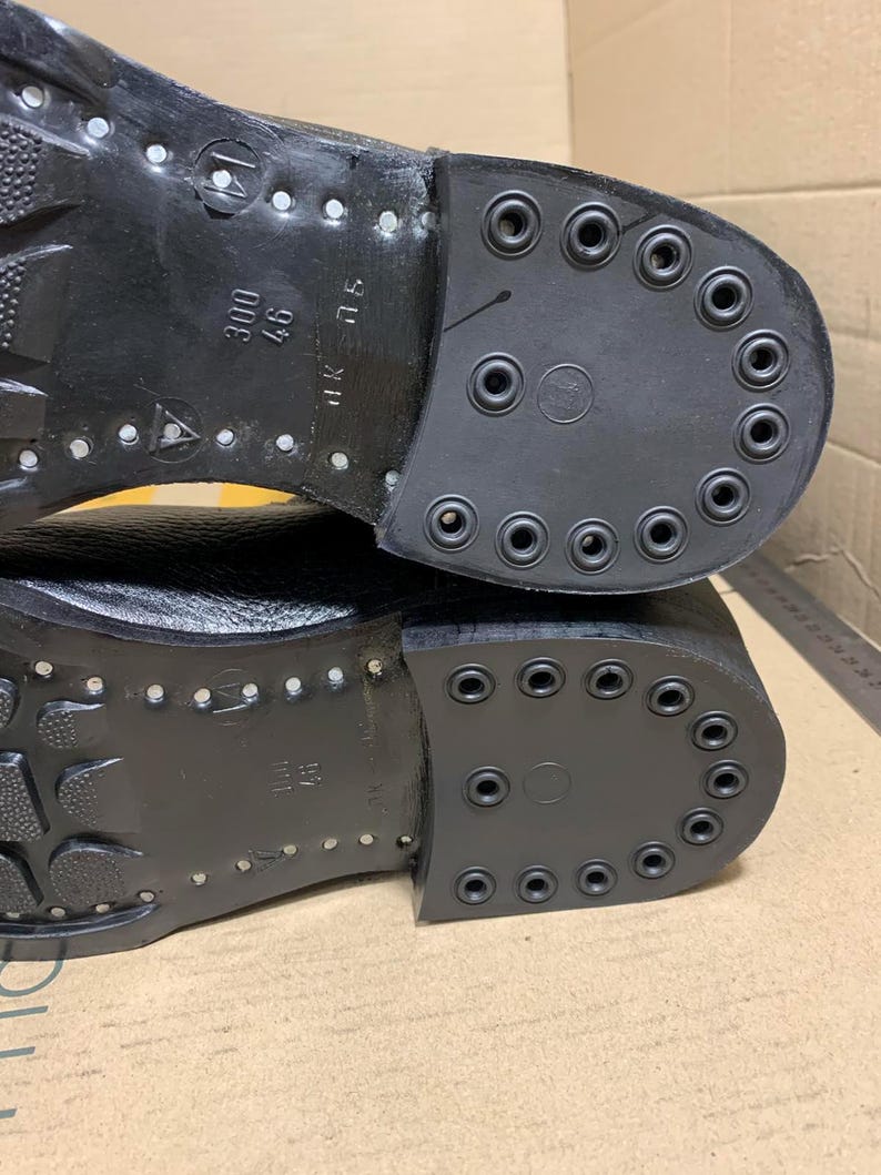 Puede incluir: Primer plano de las suelas de dos botas de cuero negro. Las suelas est&aacute;n fuertemente tachonadas con remaches met&aacute;licos y tienen un tac&oacute;n de goma grueso y duradero. Los talones tienen una serie de agujeros circulares.