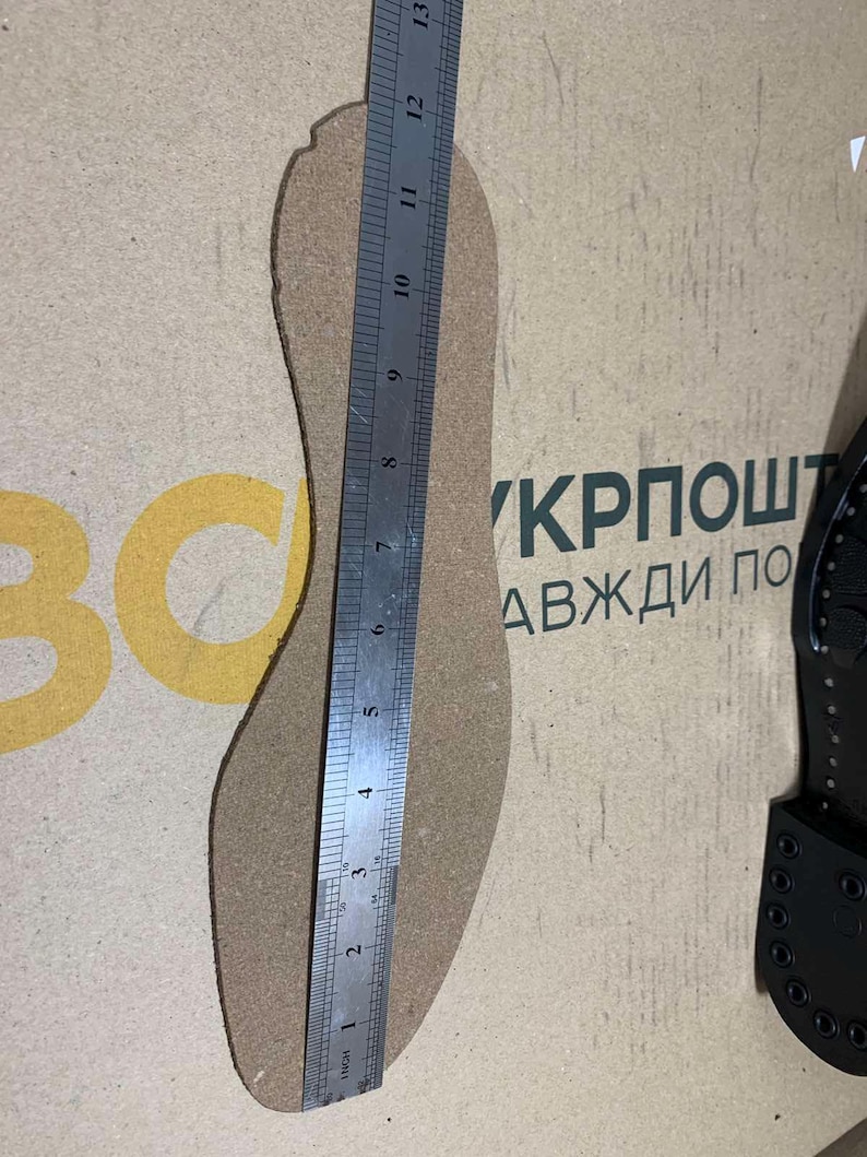 Puede incluir: Una plantilla de corcho se mide con una regla, mostrando una longitud de aproximadamente 30 cm. La plantilla es de color marr&oacute;n claro y tiene un dise&ntilde;o en forma de pie. La regla es plateada y tiene marcas en cent&iacute;metros. El fondo es una superficie marr&oacute;n claro con texto.