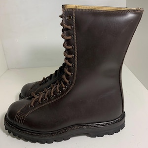 May include: A pair of brown leather boots with a tall shaft and lace-up closure. The boots have a rugged, textured sole and are designed for durability. The laces and stitching are a lighter brown, creating a contrast against the darker leather.
