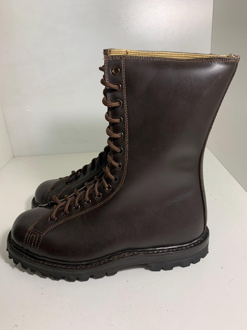 May include: A pair of brown leather boots with a tall shaft and lace-up closure. The boots have a rugged, textured sole and are designed for durability. The laces and stitching are a lighter brown, creating a contrast against the darker leather.