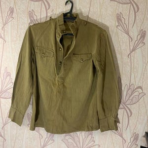 Può includere: Camicia verde oliva a maniche lunghe con colletto alla coreana e due tasche sul petto. La camicia ha chiusure con bottoni sul colletto e sui polsini. La camicia è appesa a una gruccia nera su uno sfondo a motivi floreali.