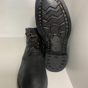 Puede incluir: Una bota de cuero negro con cordones y suela de goma texturizada con un patrón de relieve. La suela está asegurada con remaches metálicos visibles en el borde y muestra el texto 270, 42 y un logotipo grabado en la zona del talón.