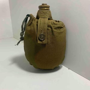 May include: An olive green canteen with a matching canvas cover. The canteen has a screw-on cap and a buttoned strap. A green drawstring secures the cover. The canteen is designed for carrying water or other liquids.