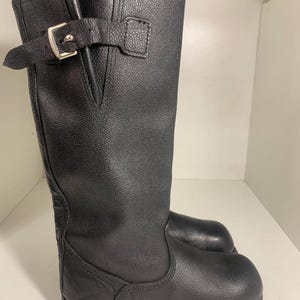 May include: Black leather knee-high boot with a buckle closure. The boot features a textured leather finish and a silver-colored buckle. The boot is designed with a rounded toe and a low heel.