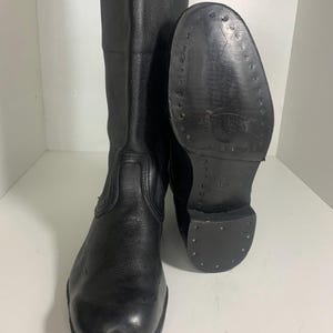 Puede incluir: Un par de botas de cuero negro. Una bota está de pie y la otra está tumbada de lado, mostrando la suela. Las suelas tienen tachuelas metálicas. Las botas son altas, llegando a la mitad de la pantorrilla.