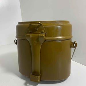 以下が含まれることがあります： オリーブグリーンの金属製メスキット。ヒンジ付きの蓋と大きなハンドルが付いています。円筒形の容器には蓋を固定するための留め金と、側面に小さなハンドルが付いています。屋外での使用を目的としています。